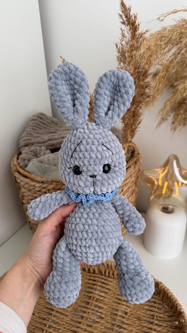 May include: A handmade crochet plush toy in the shape of a rabbit, crafted from soft grey yarn. The rabbit features long upright ears, black plastic eyes, a small stitched nose, and a light blue ruffled collar around its neck.