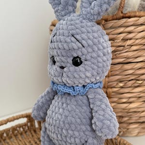 May include: A handmade crochet rabbit toy crafted from soft, plush grey yarn. The rabbit features long upright ears, black plastic safety eyes, embroidered eyebrows, and a small blue crocheted collar around its neck. The texture is thick and fuzzy.