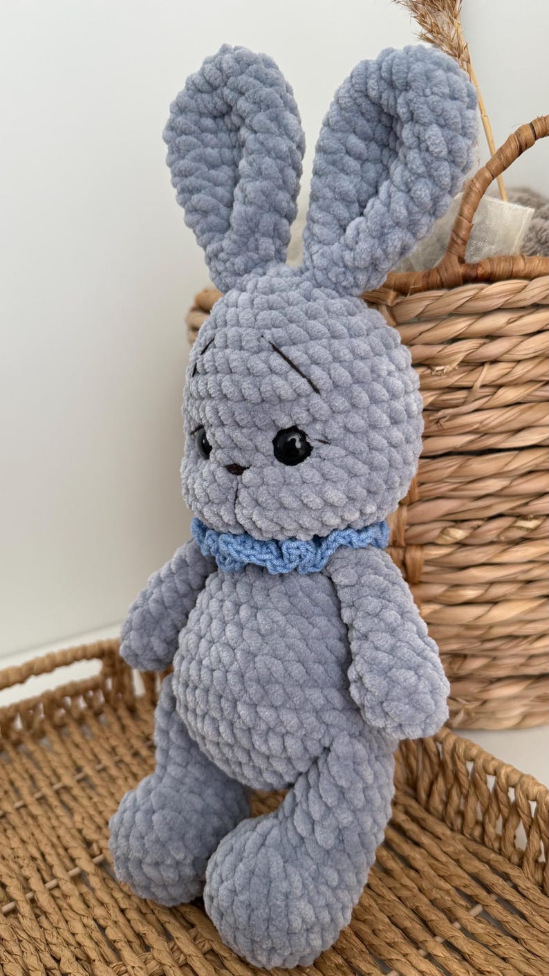 May include: A handmade crochet rabbit toy crafted from soft, plush grey yarn. The rabbit features long upright ears, black plastic safety eyes, embroidered eyebrows, and a small blue crocheted collar around its neck. The texture is thick and fuzzy.