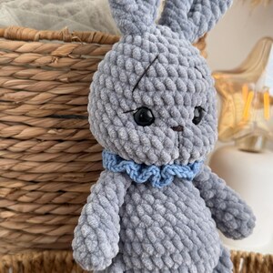 May include: A gray crocheted plush rabbit toy made from soft, chunky yarn. The rabbit features long upright ears, black plastic safety eyes, a small brown stitched nose, and a light blue ruffled collar around its neck. The texture is uniform and bumpy.
