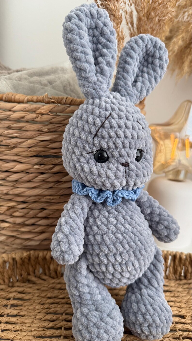 May include: A gray crocheted plush rabbit toy made from soft, chunky yarn. The rabbit features long upright ears, black plastic safety eyes, a small brown stitched nose, and a light blue ruffled collar around its neck. The texture is uniform and bumpy.