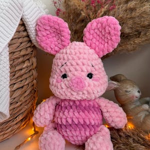 May include: A handmade crochet plush toy of a piglet with large rounded ears, a round snout, and small black eyes. The toy is crafted from soft, chunky light pink yarn, with a darker pink variegated yarn used for the round belly section.