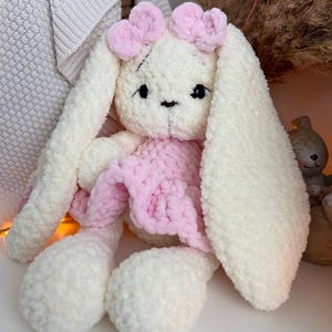 May include: A plush stuffed rabbit toy made of cream-colored yarn with long ears, featuring a pink crocheted dress and two pink bows on its head. The rabbit has embroidered black eyes and a small black nose, with a soft, textured appearance.
