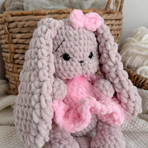May include: A plush, crocheted rabbit toy made from soft, light mauve yarn. The rabbit features long, floppy ears, black safety eyes, and embroidered facial details. It wears a small, light pink crocheted bow on its head and a pink tulle skirt.