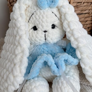 May include: A plush, white, crocheted bunny toy with long, floppy ears, black safety eyes, and embroidered facial features. The bunny wears a light blue crocheted skirt and a matching light blue bow on its head, accented by a thin, translucent white ribbon.