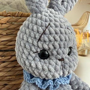May include: A gray crocheted plush rabbit toy made from soft, chunky yarn. The rabbit features long upright ears, large black plastic safety eyes, and a small brown stitched nose. A ruffled light blue yarn collar is worn around its neck.