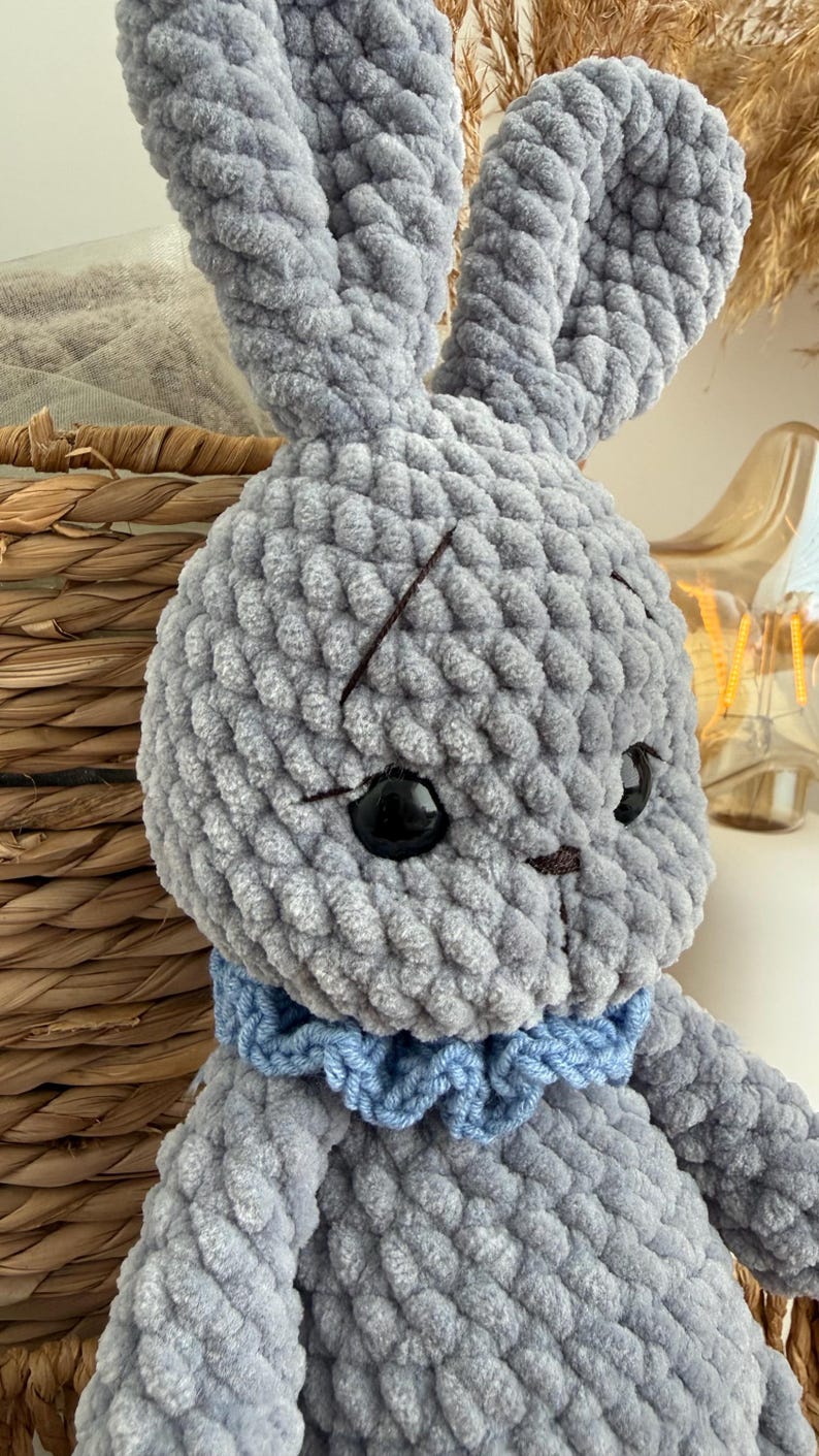 May include: A gray crocheted plush rabbit toy made from soft, chunky yarn. The rabbit features long upright ears, large black plastic safety eyes, and a small brown stitched nose. A ruffled light blue yarn collar is worn around its neck.
