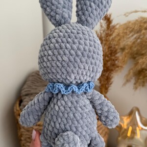 May include: A gray crocheted plush rabbit toy made from soft, textured yarn. The rabbit features long upright ears, a small round tail, and a light blue ruffled collar around its neck. The toy has a dense, uniform stitch pattern throughout its body.