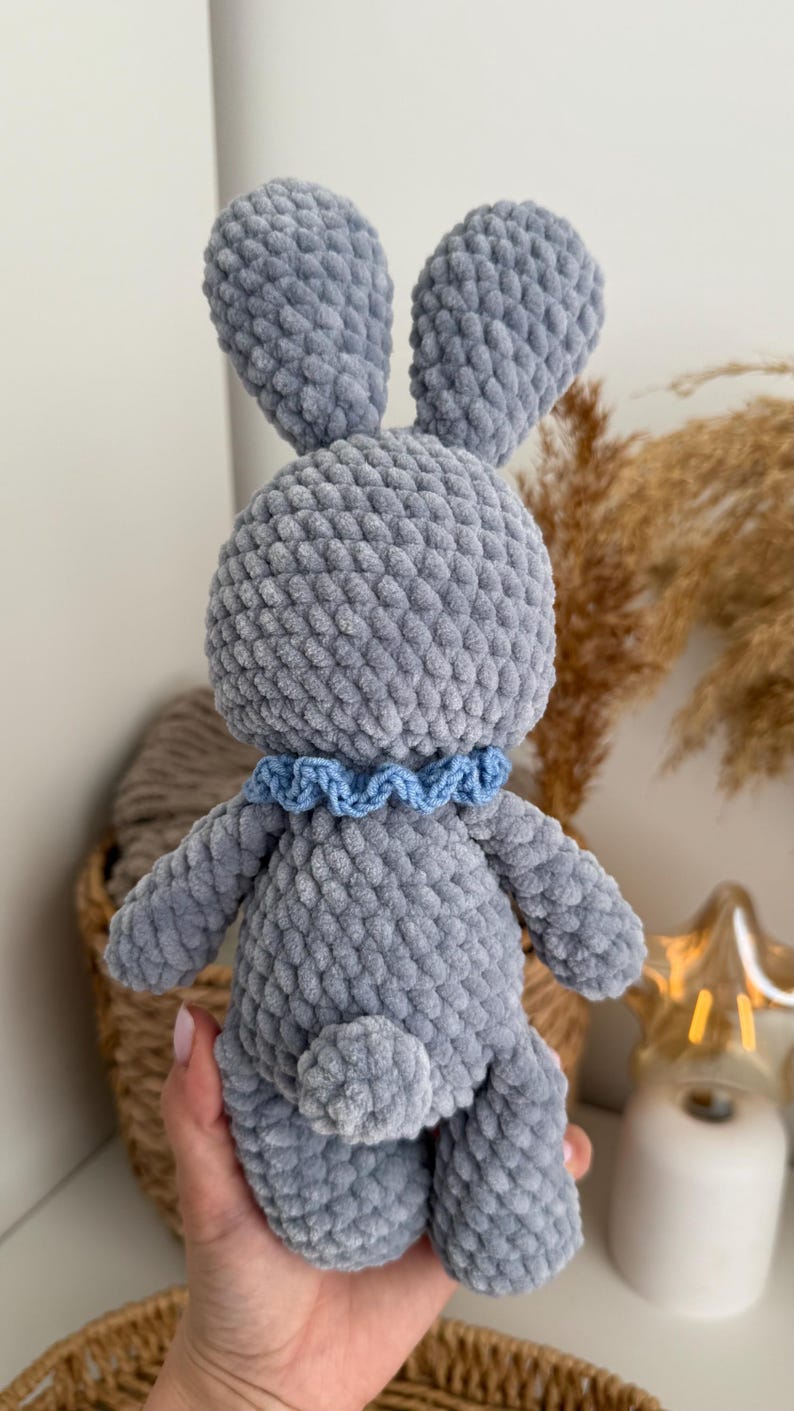 May include: A gray crocheted plush rabbit toy made from soft, textured yarn. The rabbit features long upright ears, a small round tail, and a light blue ruffled collar around its neck. The toy has a dense, uniform stitch pattern throughout its body.