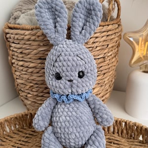 May include: A plush gray crocheted rabbit toy with long ears, black plastic eyes, and a small embroidered nose. The rabbit features a textured, soft yarn construction and wears a light blue ruffled collar around its neck, sitting upright.