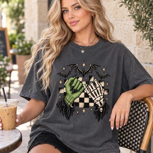 May include: A charcoal gray short-sleeve t-shirt featuring a graphic print of a green hand and a skeleton hand making a pinky promise. The design includes a black and white checkered background, several black bats, and small yellow star accents.