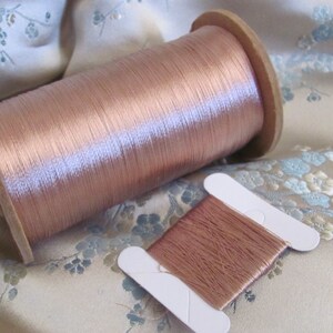 May include: A spool of rose gold colored silk thread on a wooden spool. A smaller sample of the thread is also shown.