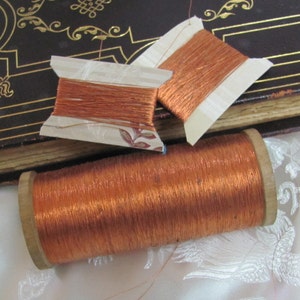 May include: Three spools of copper wire, two are on white cardboard spools and one is on a wooden spool. The spools are on a white surface with a book in the background.