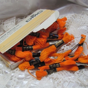 Peut inclure: Une boîte de fil à broder DMC Mouline Special 6 brins de couleur orange vif. La boîte est ouverte et le fil se répand.
