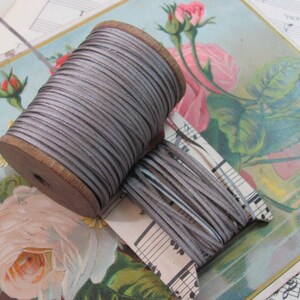May include: A spool of silver grey cord or ribbon wound around a wooden spool. The cord is displayed on a piece of sheet music with a floral design.