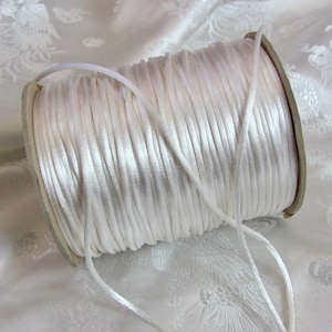 May include: A reel of white satin cord. The cord is neatly wound on a cardboard core. The cord is a brilliant white and has a smooth, shiny appearance. The background is a white fabric with a delicate pattern.