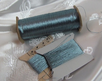 Könnte beinhalten: Zwei Spulen mit hellblauem Metallic-Garn. Eine Spule befindet sich auf einer Pappspule mit einem Notenblatt-Design. Die andere Spule befindet sich auf einer weißen Pappspule.