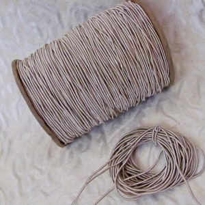 May include: A spool of beige elastic cord, wound around a cardboard tube. A smaller portion of the cord is coiled into a circle, showing the texture and color of the elastic.