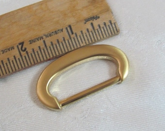 May include: A gold-colored, oval-shaped metal buckle with a straight bar closure. A wooden ruler with inch markings is in the background. The buckle is approximately 2 inches long.