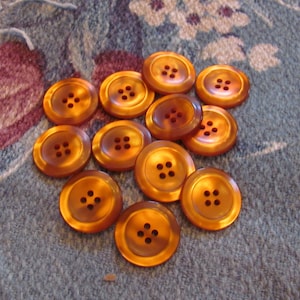 Puede incluir: Un grupo de 13 botones de color ámbar con cuatro agujeros cada uno. Los botones están dispuestos en una pila suelta sobre una superficie de tela estampada.