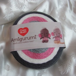 May include: A skein of Red Heart yarn in gray, white, and pink stripes. The label reads "Amigurumi" and "each one makes two". Two small crocheted elephant amigurumi toys are pictured on the label.