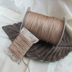 May include: A spool of beige satin cord, wound around a cardboard spool, on a wooden leaf-shaped tray. The cord is approximately 1/8 inch wide.
