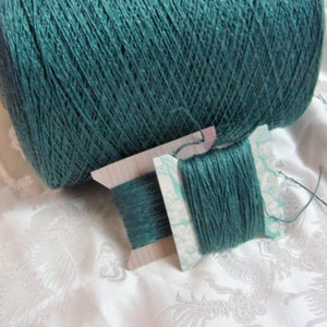 May include: A spool of teal green yarn wound around a cardboard tube. A smaller spool of the same yarn is wound on a wooden bobbin with a white paper label.