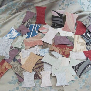 May include: A collection of paper cutouts in the shape of spools, each featuring a different patterned fabric design. The fabrics include floral, geometric, and abstract patterns in various colors like red, blue, and beige. The cutouts are scattered on a floral patterned fabric.