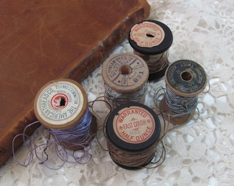 Könnte beinhalten: Vier Garnrollen, zwei mit Holzspulen und zwei mit schwarzen Spulen. Die Garnfarben sind lila, silber, braun und hellbraun. Die Spulen tragen Etiketten mit der Aufschrift "The American Thread Co.", "Tyrian Dyes", "Warranted Fast Color" und "Warranted Fast Color".