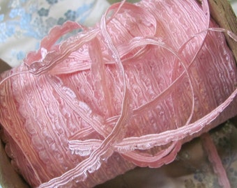 May include: A spool of pale pink ribbon with a scalloped edge. The ribbon is tightly wound on a wooden spool. The ribbon has a textured appearance and is likely made of fabric. The background is a floral patterned fabric.
