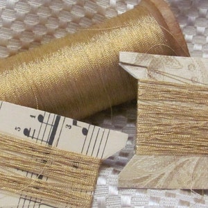 May include: Two spools of gold metallic thread. One spool is on a wooden bobbin and the other is on a cardboard spool. The cardboard spool has a music sheet design.