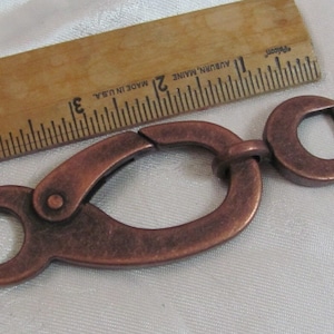 May include: A copper-toned metal swivel clasp with a rounded oval shape and a smaller, round opening. The clasp is approximately 2 inches long.