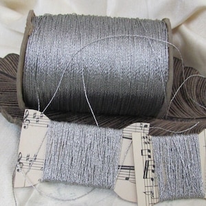 Może przedstawiać: Dwa szpulki srebrnej nici metalicznej, jedna na drewnianej szpulce i dwie na kartonowych szpulkach z wzorami nut muzycznych.