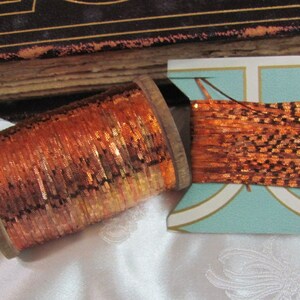 May include: Two spools of copper-coloured metallic thread. One spool is on a wooden bobbin and the other is on a white and blue cardboard spool.