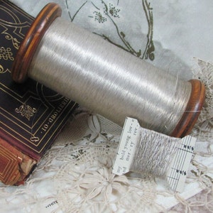 May include: A spool of silver thread with a wooden bobbin, resting on a vintage book and lace fabric. The thread is labeled "my bold young love - mer - ry rev."