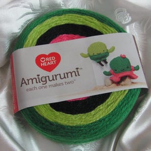 Op de afbeelding: Een bol garen in de kleuren groen, limoengroen, zwart en roze. Het garen is gelabeld "Red Heart Amigurumi" en er staat "elke bol maakt twee projecten".
