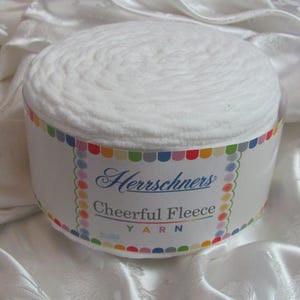 Puede incluir: Un rollo blanco de hilo de vellón alegre de Herrschners. El hilo está envuelto en una etiqueta colorida con el nombre de la marca y la descripción del producto.