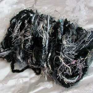 May include: A bundle of black, white, and silver yarn with a fuzzy texture. The yarn is arranged in a loose, messy pile.