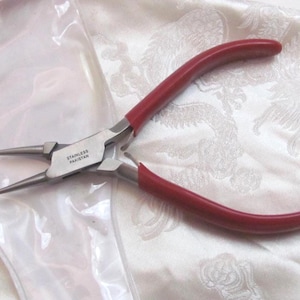 Puede incluir: Alicates de punta fina con mango rojo y cabeza de metal plateado. Los alicates están etiquetados como "Stainless Pakistan".