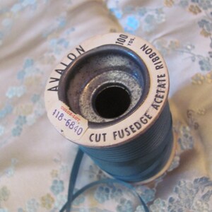 K&ouml;nnte beinhalten: Eine Spule mit tealblauem Acetatband mit einem wei&szlig;en Etikett, auf dem "Avalon Cut Fusedge Acetate Ribbon 100 Yds. No. 888 Color-Lot 118-6840" steht.