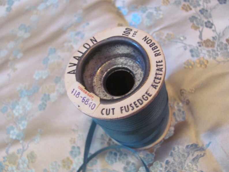 K&ouml;nnte beinhalten: Eine Spule mit tealblauem Acetatband mit einem wei&szlig;en Etikett, auf dem "Avalon Cut Fusedge Acetate Ribbon 100 Yds. No. 888 Color-Lot 118-6840" steht.
