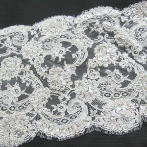 May include: Close-up of white lace fabric with floral and scrollwork patterns. The lace is embellished with sequins and small, round beads. The fabric has a scalloped edge and is likely used for bridal or formal wear.