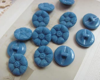 May include: A collection of 11 blue plastic buttons with a variety of designs. Some buttons have a floral pattern, while others have a simple four-hole design.