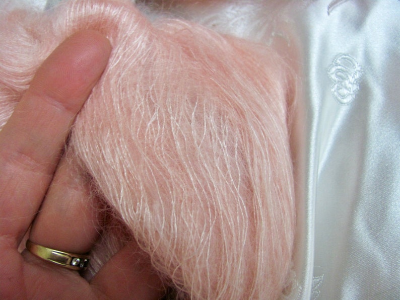 K&ouml;nnte beinhalten: Nahaufnahme eines weichen, hellrosa Garns mit seidenartiger Textur. Das Garn wird in einer Hand gehalten, und der Hintergrund ist ein wei&szlig;er Satin-Stoff.