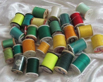 May include: A collection of spools of thread in various colors, including green, yellow, orange, red, and white. The spools are arranged in a pile on a white surface.