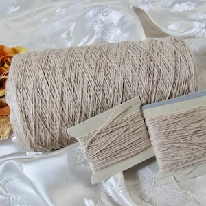 May include: A large cylindrical spool of light beige yarn positioned next to two smaller rectangular cardboard bobbins wrapped in the same light beige yarn, accompanied by a cluster of orange and yellow fabric flower petals.