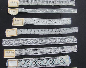May include: Collection of white lace trims with various delicate patterns, including floral and geometric designs. Each trim is labeled with a small tag containing numbers. The trims are displayed against a dark background, showcasing the intricate details of the lace.