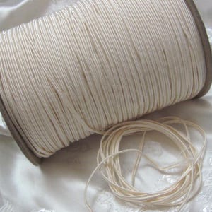 May include: A large spool of cream-coloured cord, tightly wound, with a small pile of loose cord in the foreground. The cord is a neutral colour, suitable for various crafting projects. The spool has brown edges.