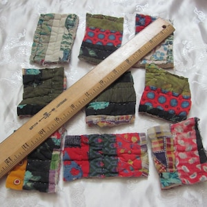 K&ouml;nnte beinhalten: Neun Stoffquadrate mit Patchwork-Muster in verschiedenen Farben, darunter Rot, Gr&uuml;n, Blau und Wei&szlig;. Die Quadrate sind in einem zuf&auml;lligen Muster auf einer wei&szlig;en Oberfl&auml;che angeordnet. Ein Holzliniall liegt auf den Quadraten.
