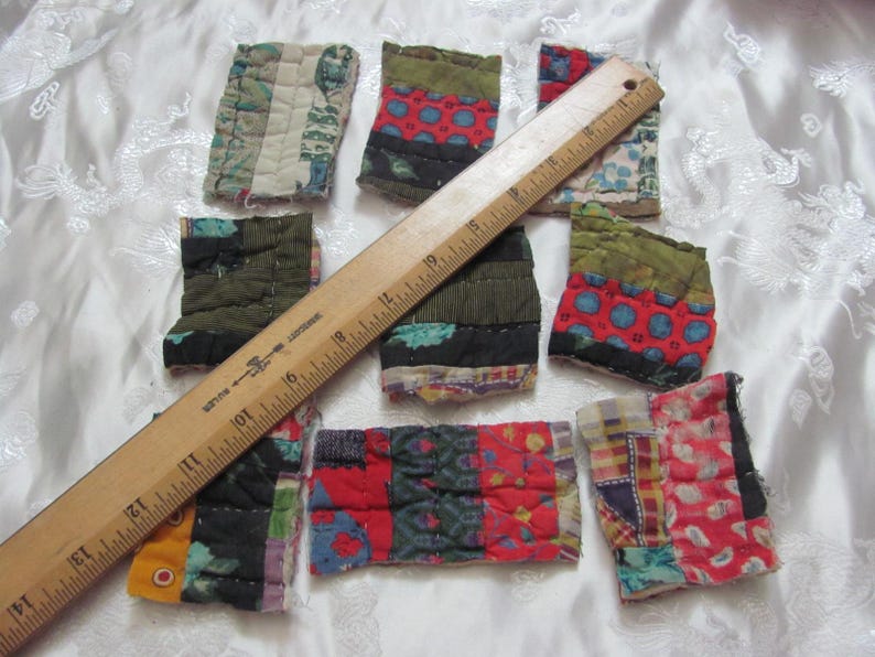 K&ouml;nnte beinhalten: Neun Stoffquadrate mit Patchwork-Muster in verschiedenen Farben, darunter Rot, Gr&uuml;n, Blau und Wei&szlig;. Die Quadrate sind in einem zuf&auml;lligen Muster auf einer wei&szlig;en Oberfl&auml;che angeordnet. Ein Holzliniall liegt auf den Quadraten.